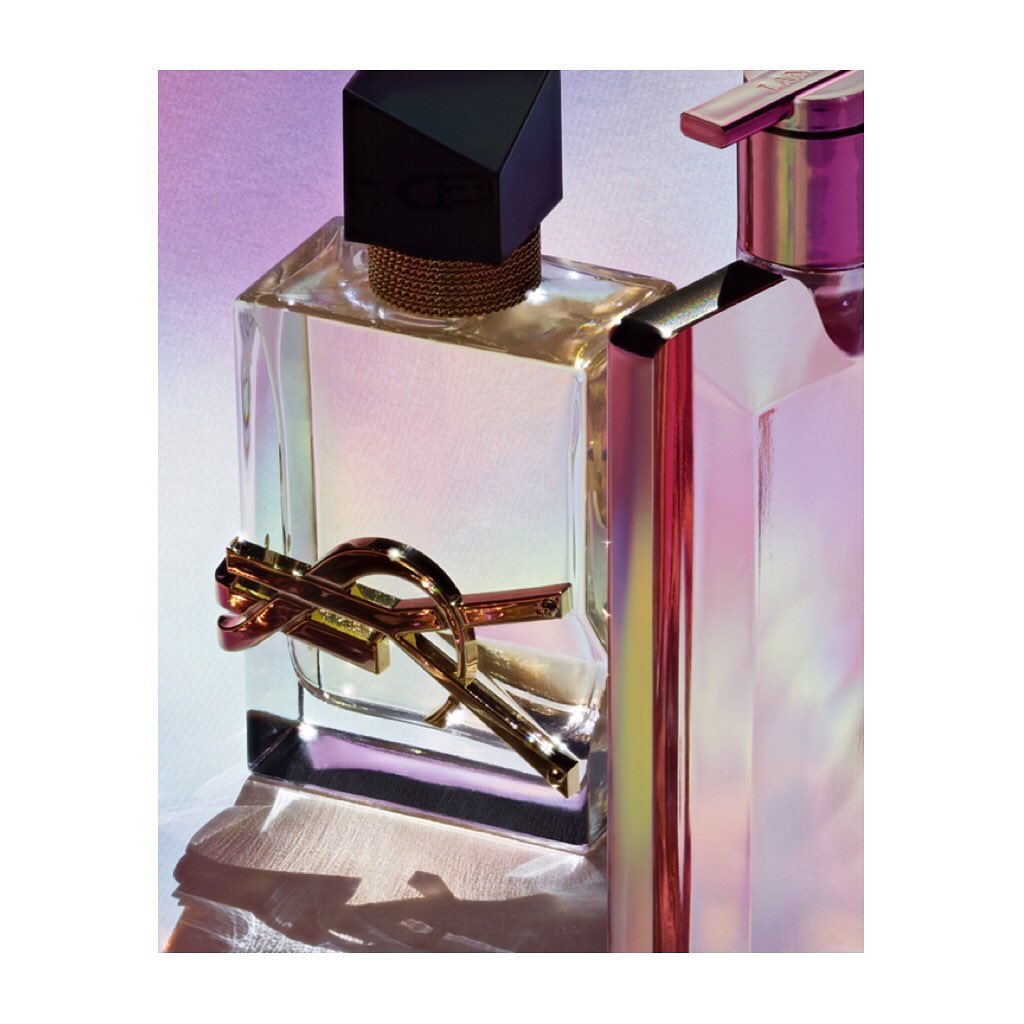 Two elegant glass perfume bottles reflect light, with a YSL logo prominently displayed on one, set against an iridescent background.
