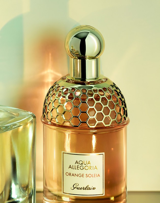 Close-up studio shot of Aqua Allegoria Orange Soleia Guerlain perfume bottle with a gold honeycomb pattern on top against a soft yellow background.