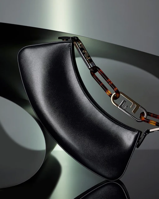 Studio shot of a black leather Fendi handbag with tortoise shell-colored chain strap, displayed against a reflective surface and background.