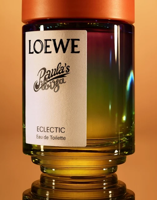 Close-up studio shot of Loewe Paula's Ibiza Eclectic Eau de Toilette glass bottle with orange cap, set on a reflective surface.