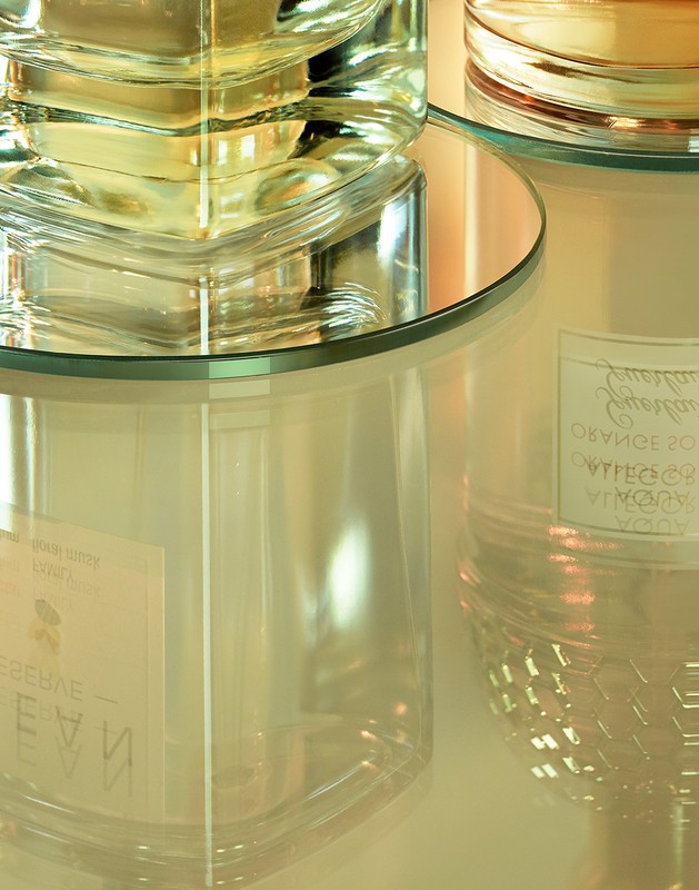 Close-up of transparent glass perfume bottle and candle jar on a reflective surface, casting soft reflections and highlights in a studio setting.