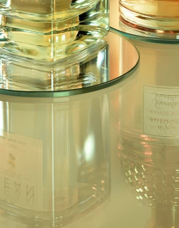 Close-up of transparent glass perfume bottle and candle jar on a reflective surface, casting soft reflections and highlights in a studio setting.