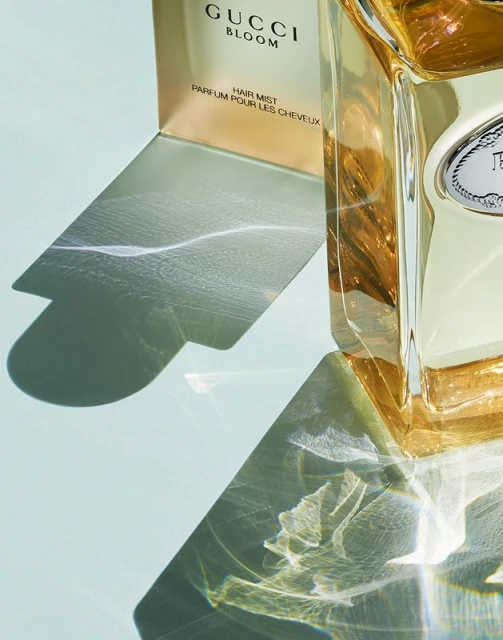 Close-up of a Gucci Bloom Hair Mist bottle with its box casting shadows on a light blue surface, creating a soft, diffused lighting effect.