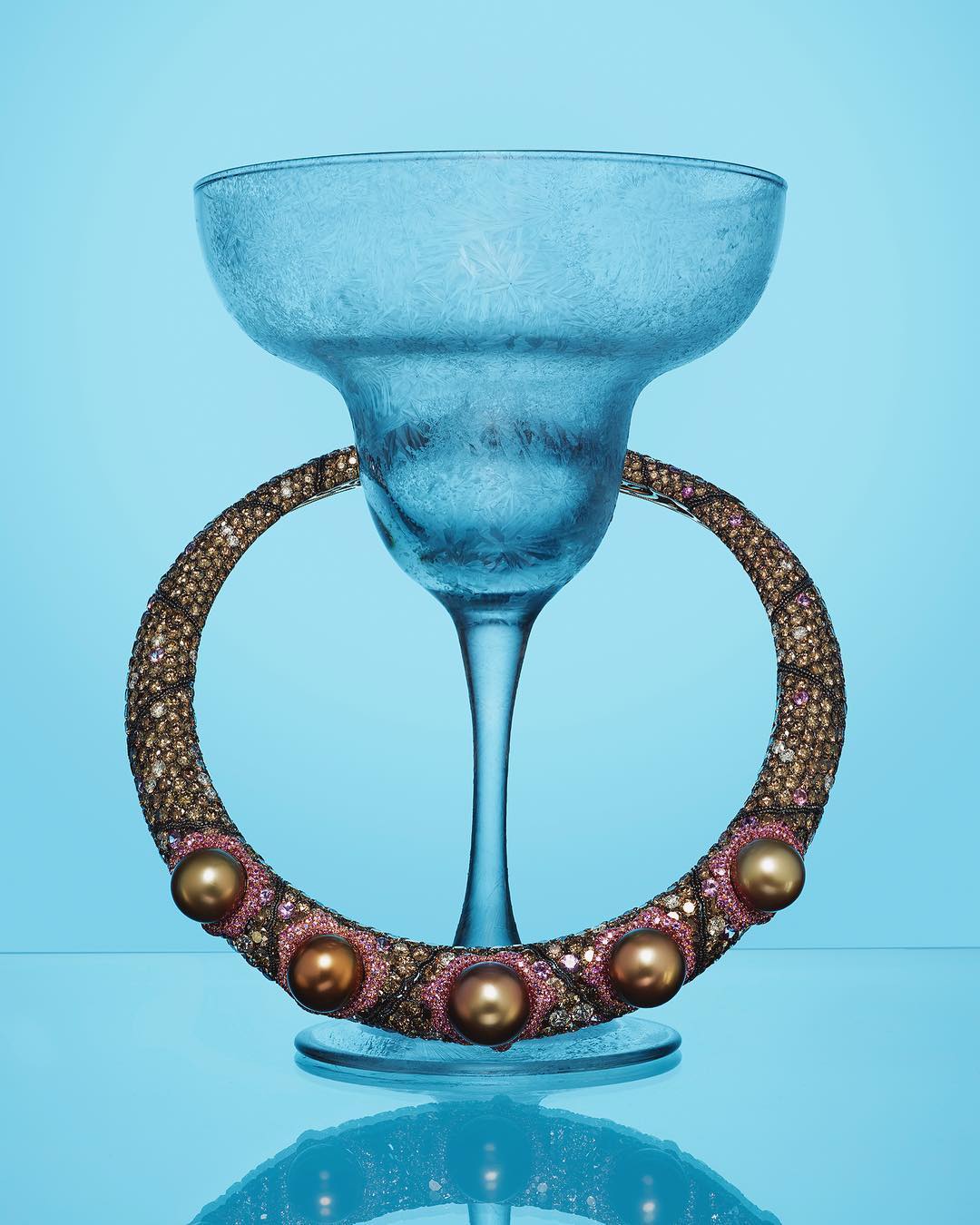 Still life shot of a margarita glass with frosted texture and an embellished pearl necklace on a graduated blue background.