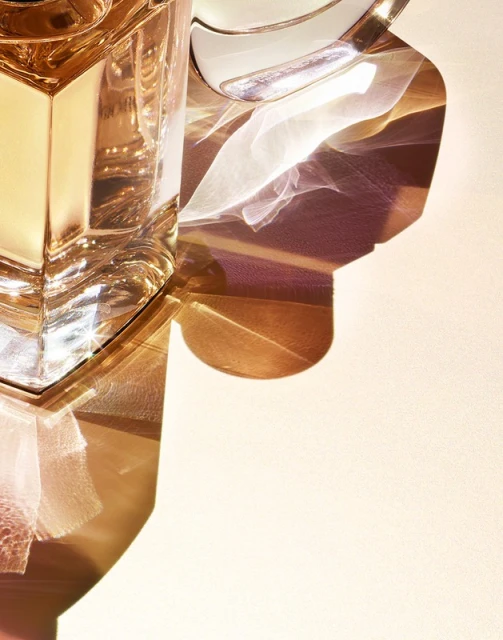 Close-up of a clear glass perfume bottle with a metallic top, casting light and shadow on a pale surface, creating a warm ambiance.