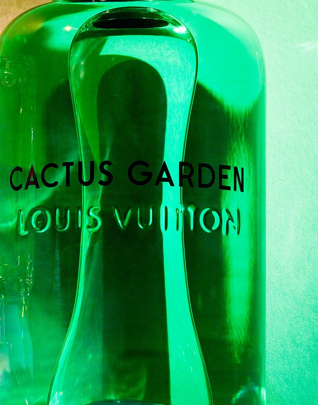 Close-up of two green-tinted glass bottles, featuring the black text 'Cactus Garden' and 'Louis Vuitton' against a soft green backdrop.