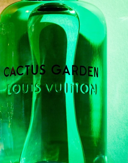 Close-up of two green-tinted glass bottles, featuring the black text 'Cactus Garden' and 'Louis Vuitton' against a soft green backdrop.