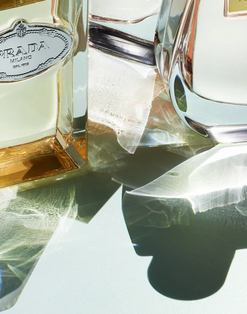 Two perfume bottles, one square and one round, with refracted light patterns against a soft blue background, showcasing minimalist design.
