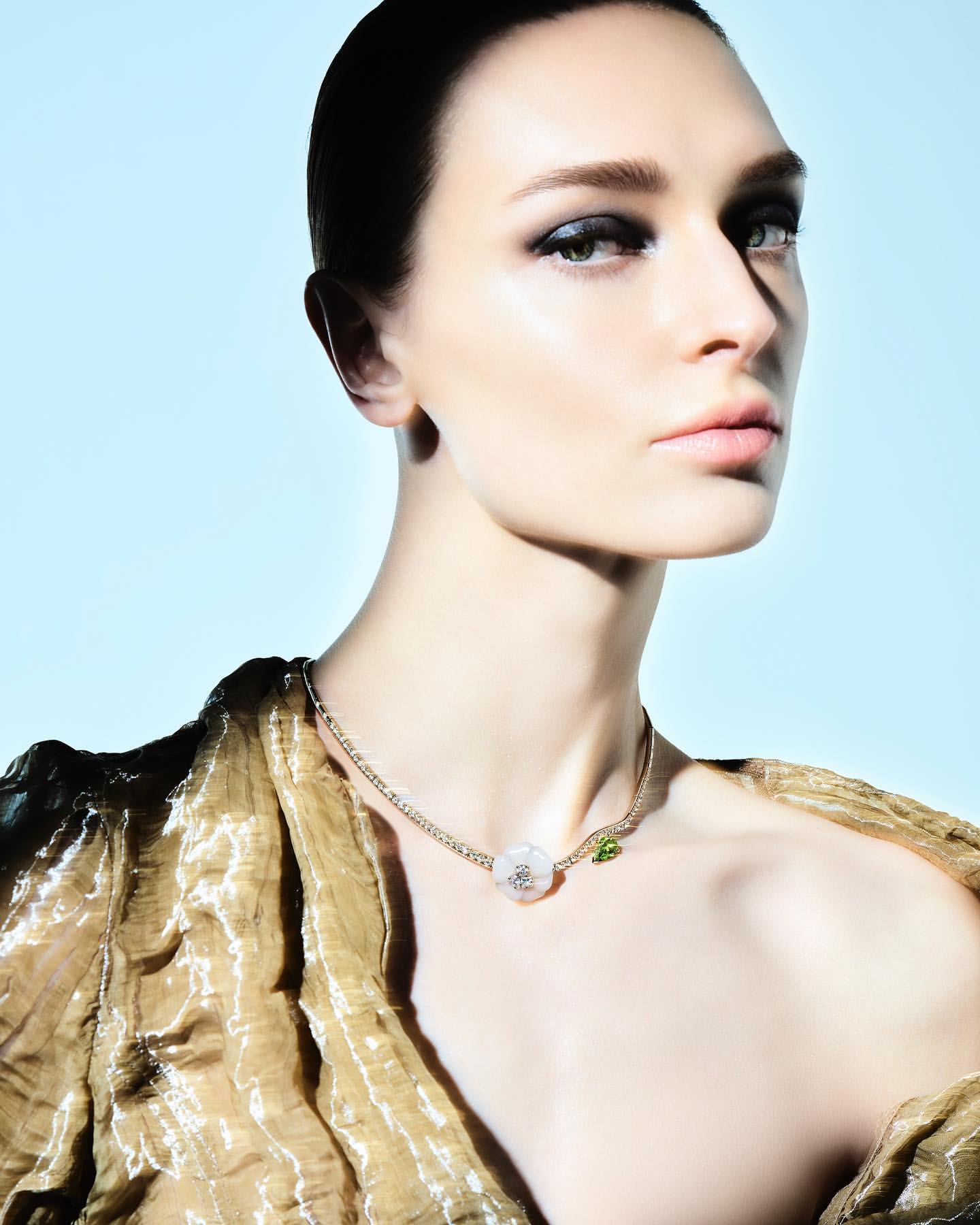 Close-up portrait of a woman wearing a delicate gold pendant necklace and a textured gold top against a pale blue background.