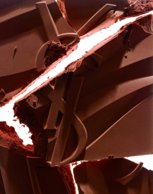 Macro shot of broken brown powder with embossed YSL logo and bright white crevices highlighting the makeup texture and color.