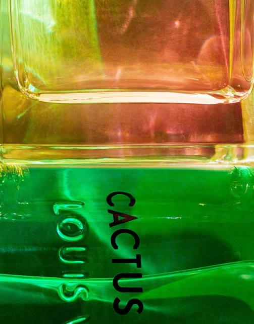Close-up of a transparent, rectangular glass bottle with gradient green and orange liquid. 'Louis Vuitton' and 'Cactus' text visible.