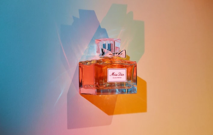 A glass perfume bottle of Miss Dior Eau de Parfum with a bow, set against a colorful wall with light and shadow gradients.