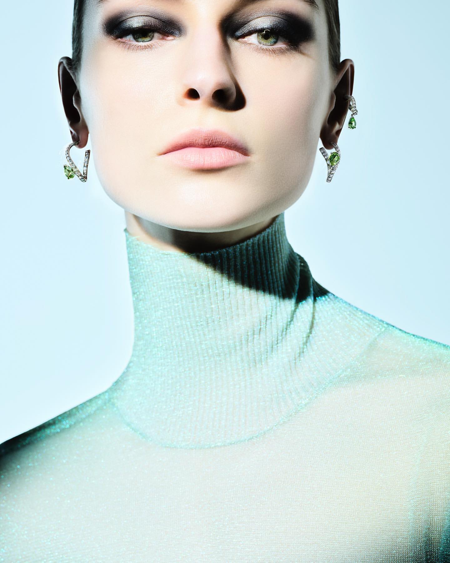 Close-up portrait of a woman with dramatic eye makeup, wearing green stone earrings and a shimmering turtleneck against a blue background.