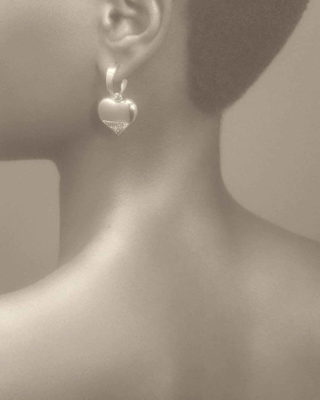 Heart shaped earrings on a person, against a neutral backdrop. The earrings are silver and hang below the ear. The person has fair skin.