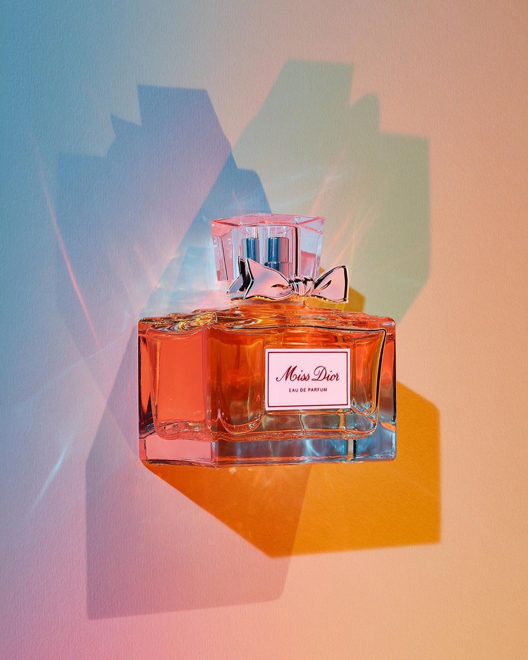 Elegant Miss Dior perfume bottle with faceted cap and bow, refracted rainbow light creating colorful shadows on a plain background.