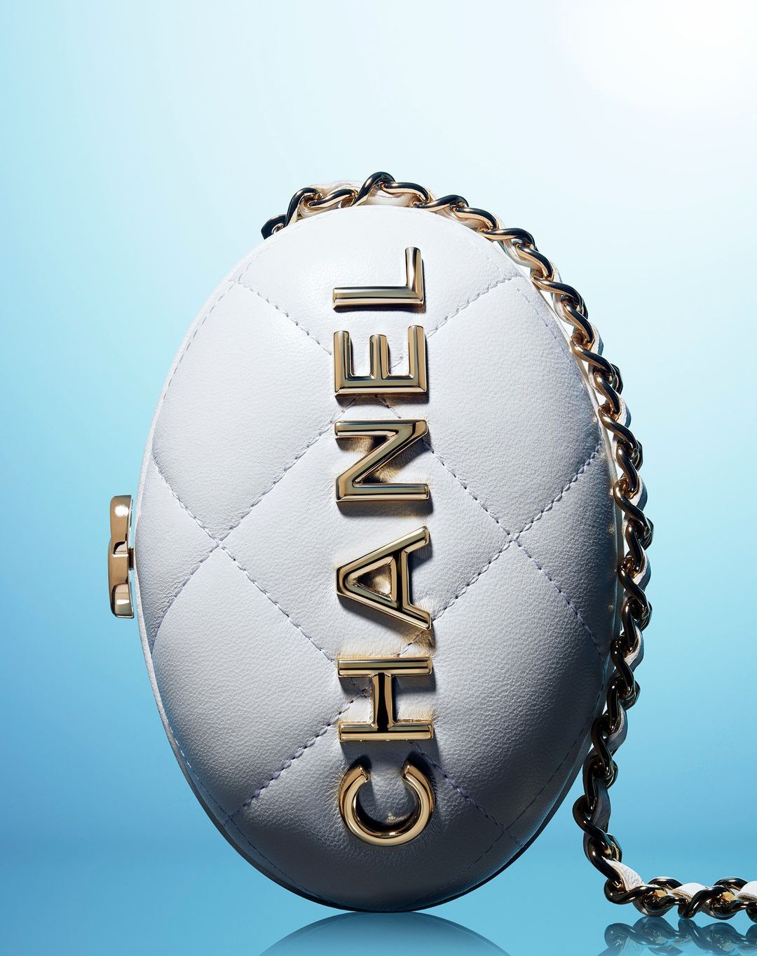 Elegant Chanel handbag in an egg-shaped design, featuring a white quilted leather, gold chain strap, and gold logo lettering against blue background.
