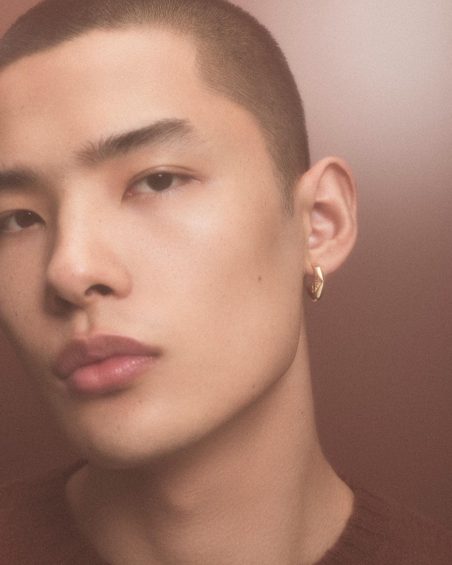 Close-up portrait of a young Asian man with short hair and gold hoop earring, wearing a dark brown sweater in a warm-toned studio.