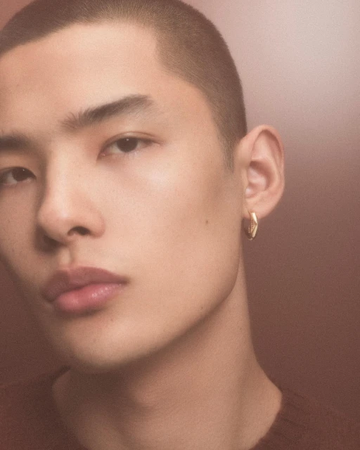Close-up portrait of a young Asian man with short hair and gold hoop earring, wearing a dark brown sweater in a warm-toned studio.