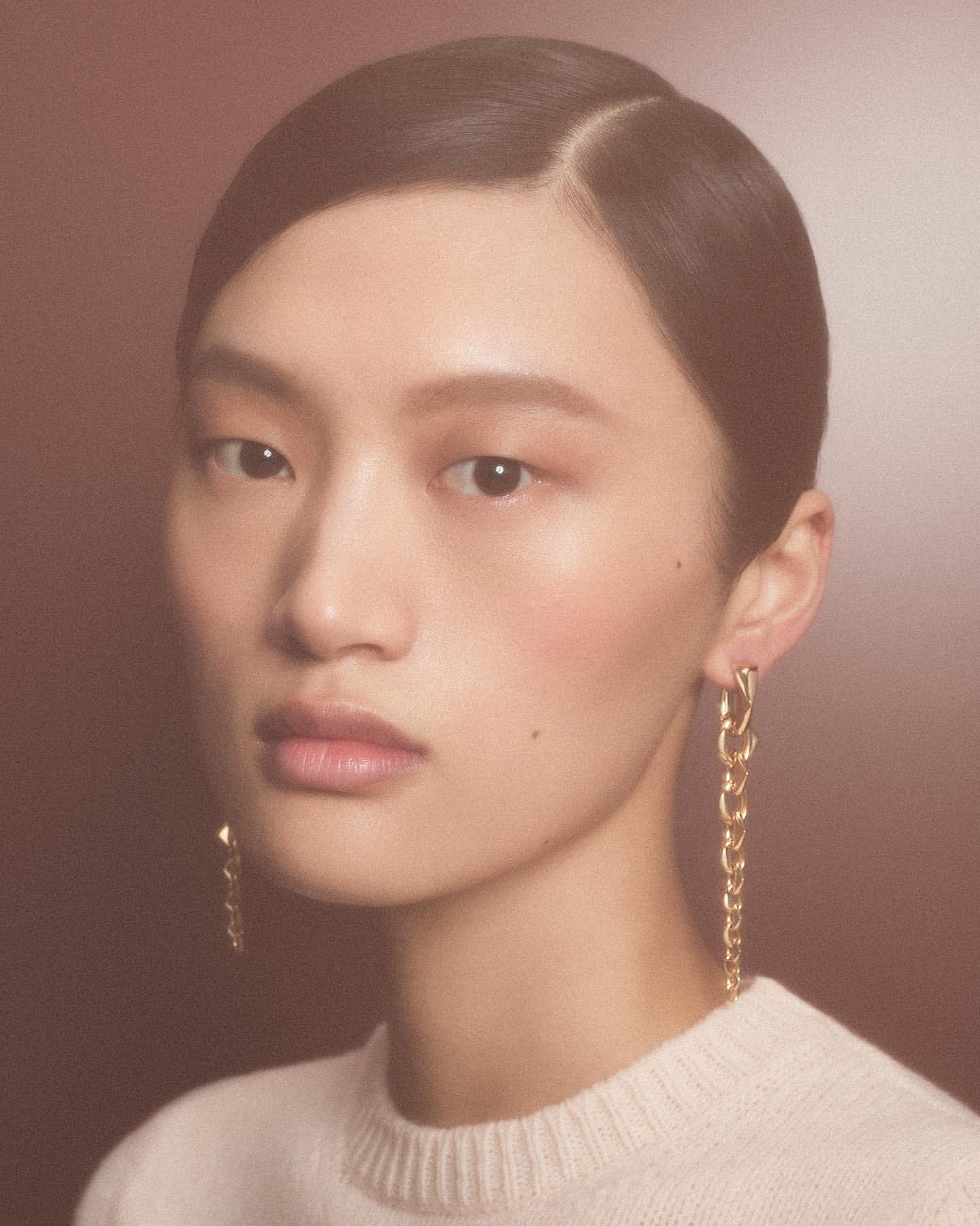 Close-up portrait of an Asian woman with sleek dark hair and gold chain earrings wearing a cream sweater against a gradient brown background.