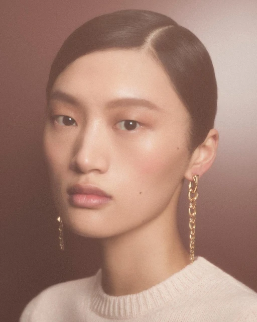 Close-up portrait of an Asian woman with sleek dark hair and gold chain earrings wearing a cream sweater against a gradient brown background.