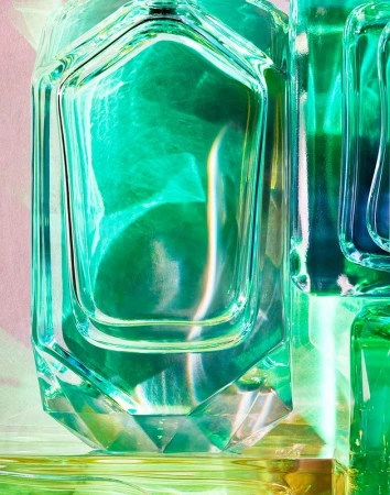 Close-up of geometric translucent green glass perfume bottles reflecting light with a pink and yellow background