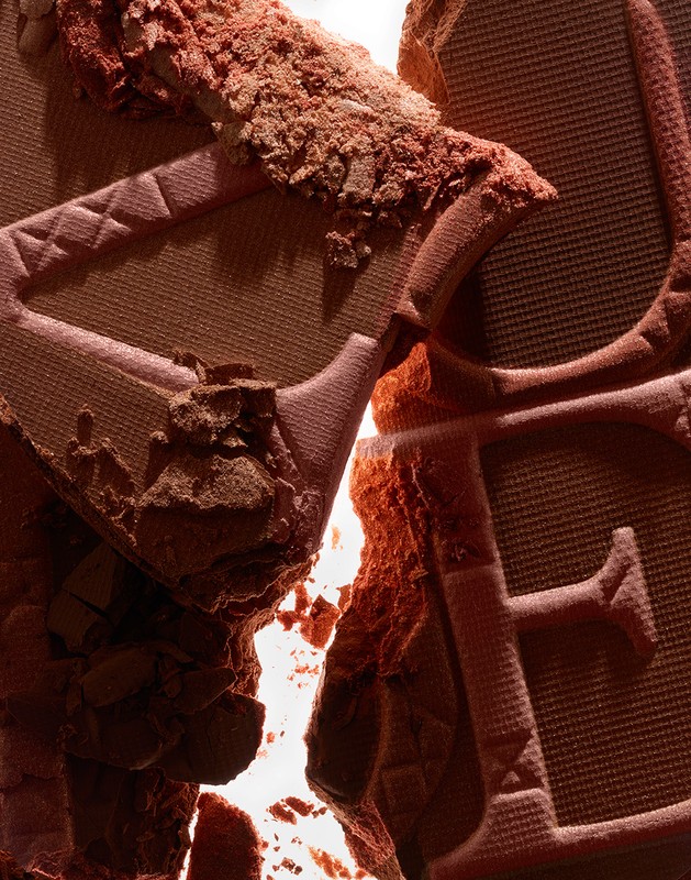 Close-up shot of broken brown cosmetic powder with a visible embossed logo, creating a textured, abstract beauty product image.