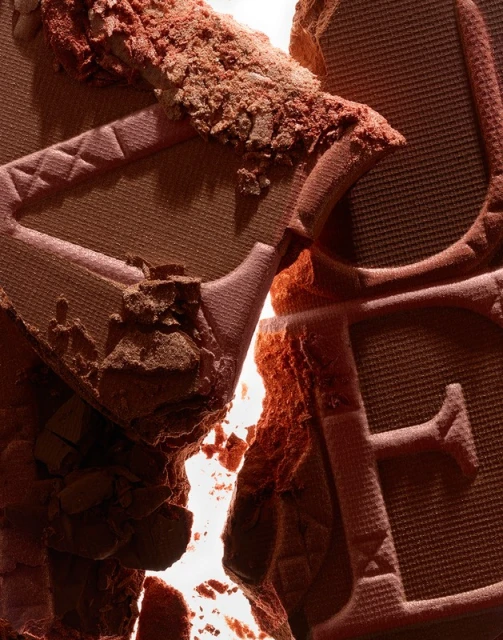 Close-up shot of broken brown cosmetic powder with a visible embossed logo, creating a textured, abstract beauty product image.