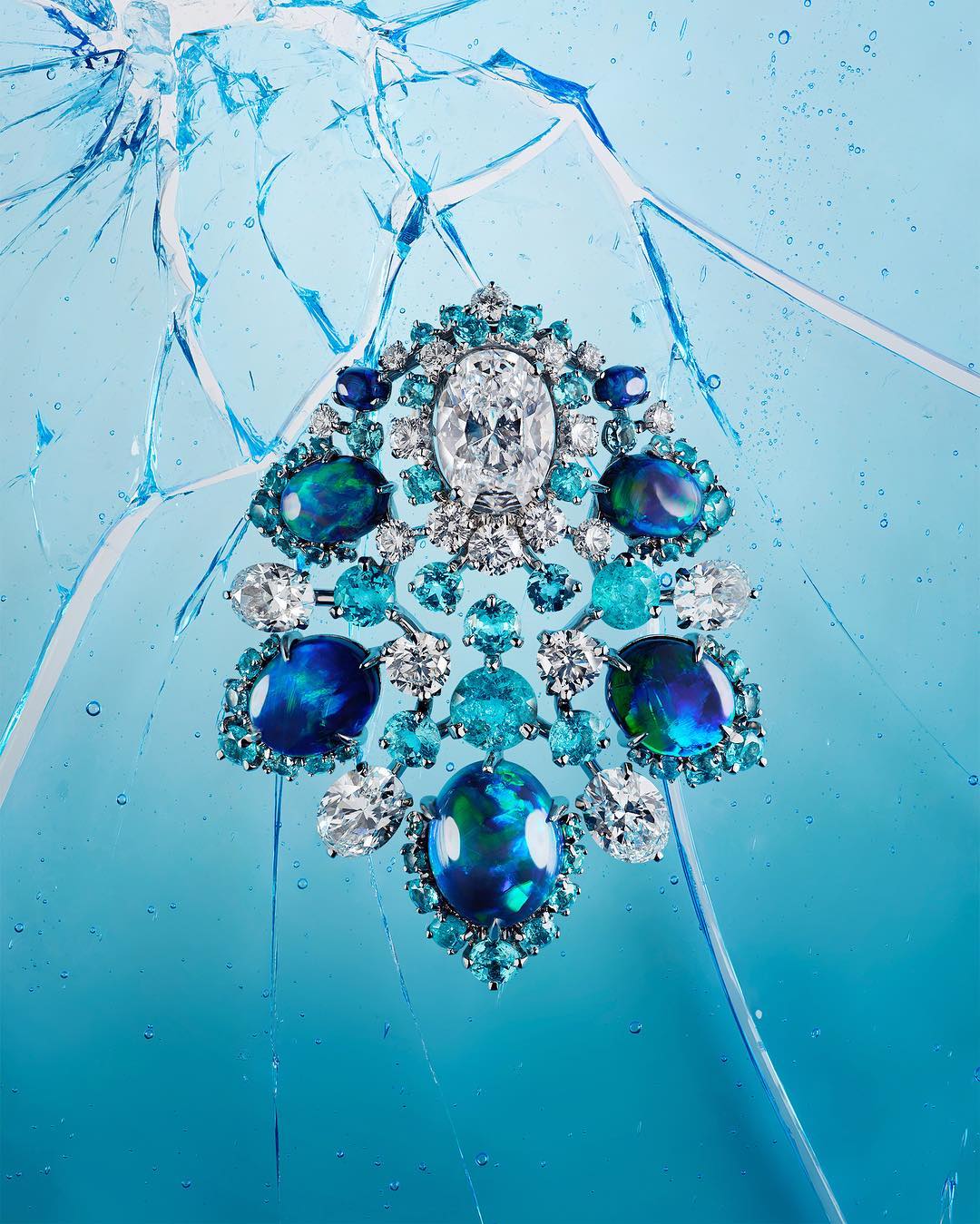 Opulent jeweled brooch on a light blue background with simulated cracked glass. The brooch features white, blue, and green stones.