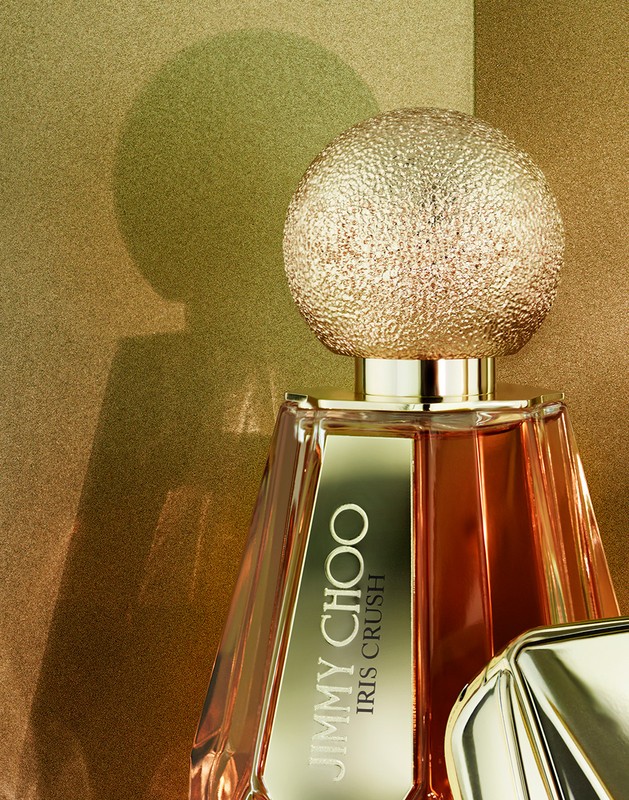 Close-up studio shot of a bottle of Jimmy Choo Iris Crush perfume with a textured, golden spherical cap against a golden background.