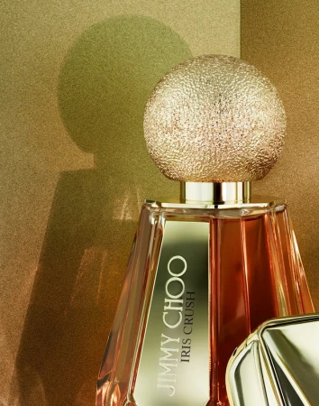 Close-up studio shot of a bottle of Jimmy Choo Iris Crush perfume with a textured, golden spherical cap against a golden background.