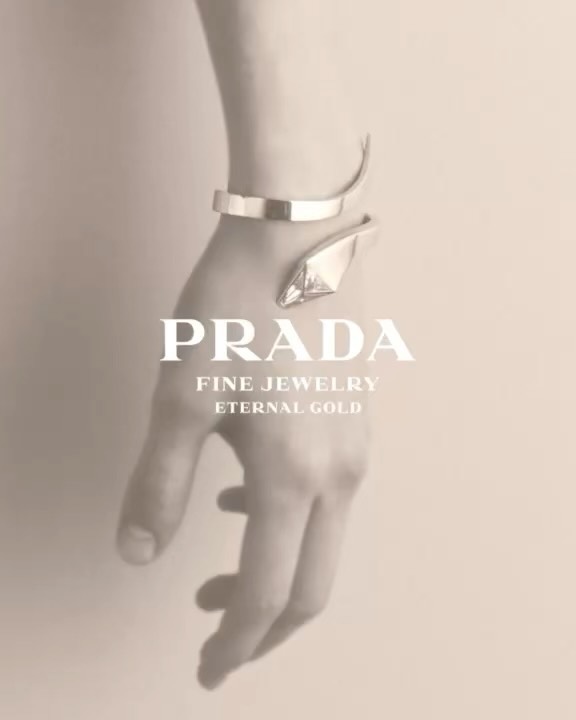 Close-up of a person's arm wearing a gold Prada bracelet against a neutral background with logo text. Fashion advertisement.