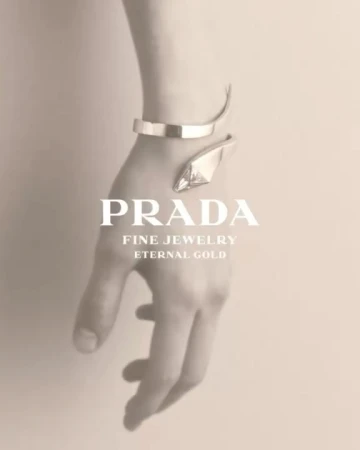 Close-up of a person's arm wearing a gold Prada bracelet against a neutral background with logo text. Fashion advertisement.