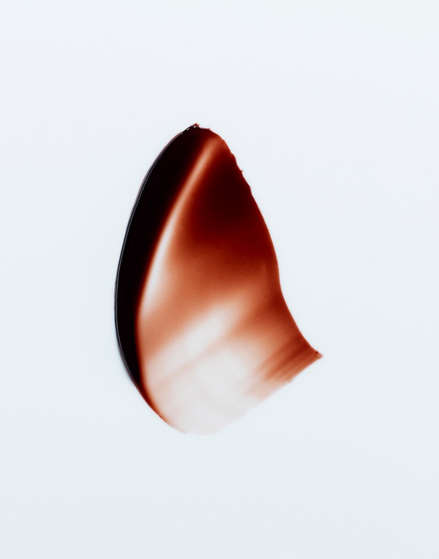 Close-up studio shot of a smear of dark brown cosmetic product, set against a clean white backdrop. Rich textures and color contrast.
