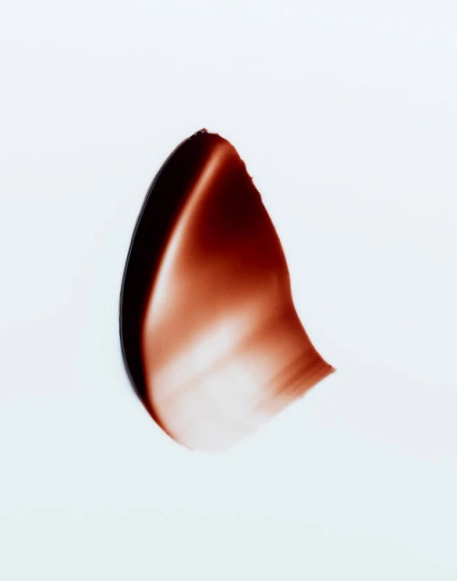 Close-up studio shot of a smear of dark brown cosmetic product, set against a clean white backdrop. Rich textures and color contrast.