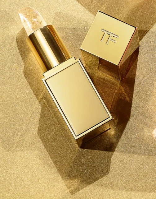 Close-up of an open gold lipstick tube with clear balm against a textured gold background, with the lipstick cap set off to the side.