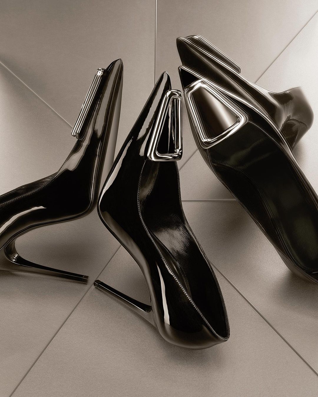 Studio shot of three black patent leather high heel shoes with silver hardware accents on a glossy tile surface, fashion forward footwear.