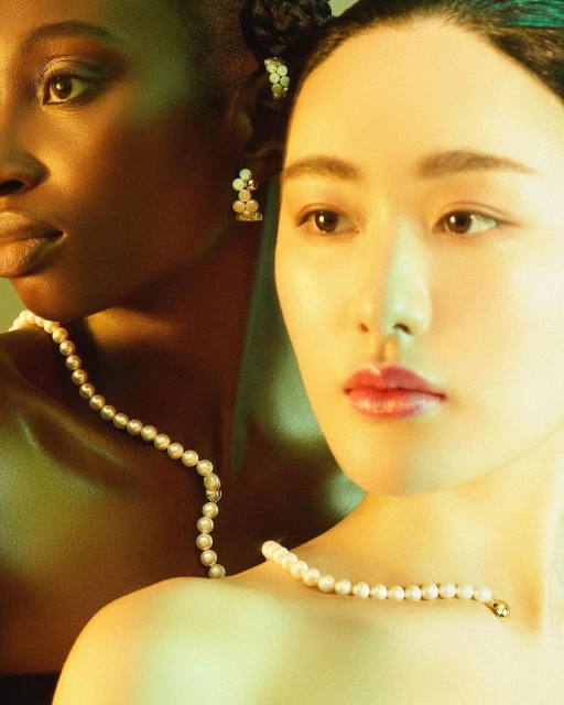 Portrait of two women with fair and dark skin wearing pearl necklaces and pearl earrings posing in a yellow-toned lit photo studio.