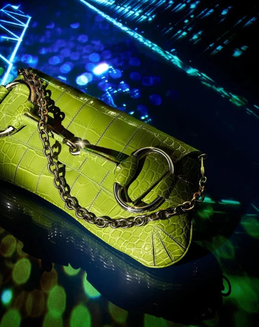 Close-up of a green faux crocodile handbag with chain detail, resting on a reflective surface, with a blurred blue and green background.