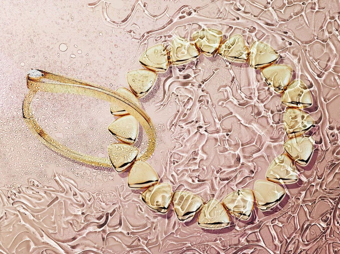 Overhead shot of a gold bracelet and necklace arranged on a reflective pink surface with rippling water-like textures and droplets.