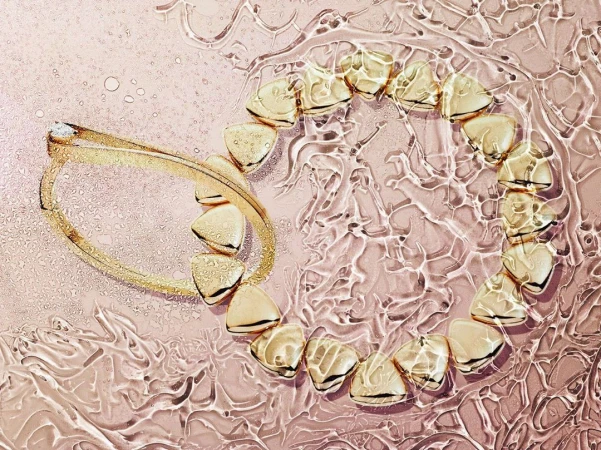 Overhead shot of a gold bracelet and necklace arranged on a reflective pink surface with rippling water-like textures and droplets.