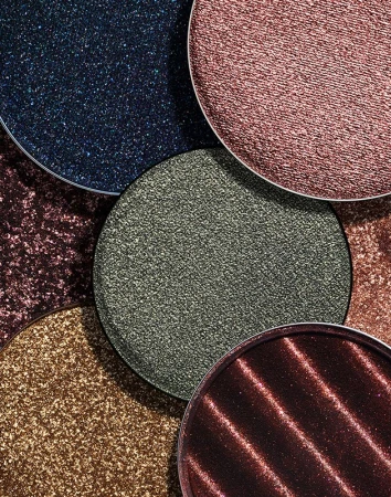 Close-up of shimmery eyeshadow palettes in blue, brown, green, pink, and copper, creating a textured, colorful display of beauty products.