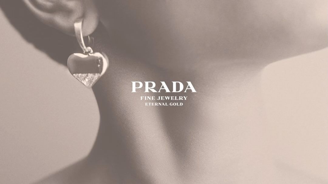Closeup of a woman's ear wearing a heart-shaped Prada earring, with a focus on jewelry design and fine gold detailing on a beige background.