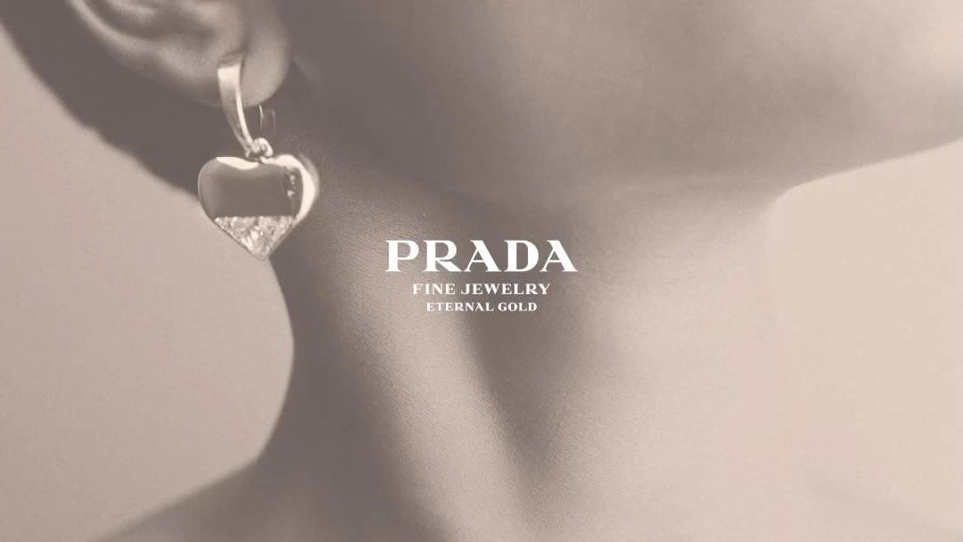 Closeup of a woman's ear wearing a heart-shaped Prada earring, with a focus on jewelry design and fine gold detailing on a beige background.
