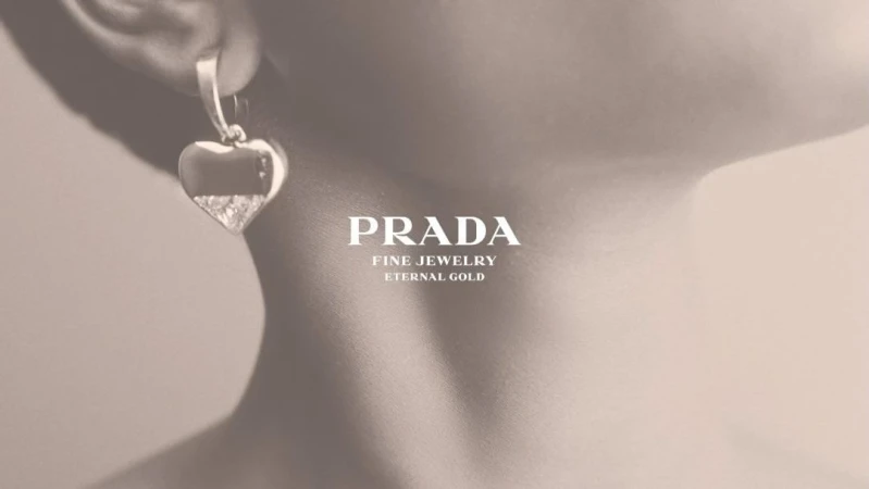 Closeup of a woman's ear wearing a heart-shaped Prada earring, with a focus on jewelry design and fine gold detailing on a beige background.