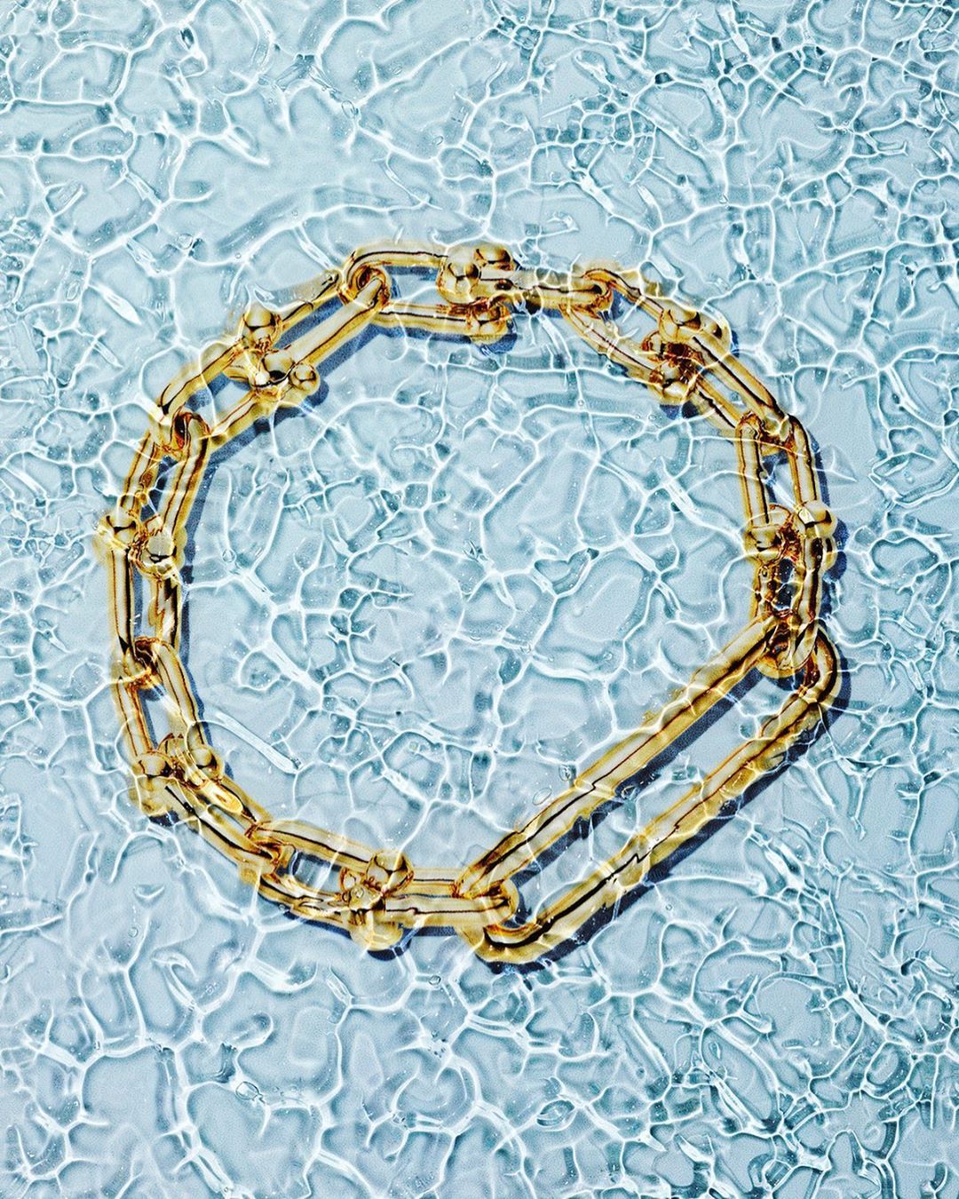Golden chain necklace in clear water with light blue hue and rippling pattern, jewelry advertisement or fashion editorial.