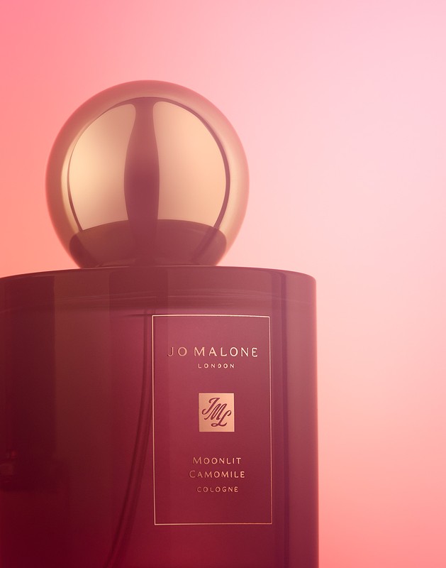 Jo Malone London Moonlit Camomile Cologne bottle with gold spherical cap against a gradient pink background in a studio shot.