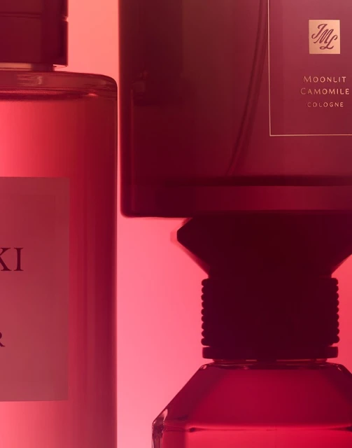 Two perfume bottles, one with a red label and one with a black label, are displayed against a gradient red backdrop in a studio setting.
