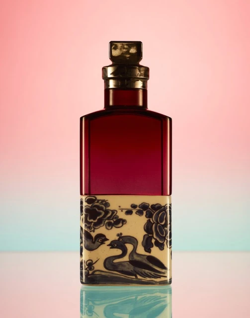 Elegant red glass bottle with a patterned lower half featuring swan and floral designs, set against a soft pink to blue gradient backdrop.