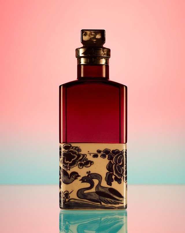 Elegant red glass perfume bottle with a gold top and decorative white floral pattern on a gradient pink and blue background.