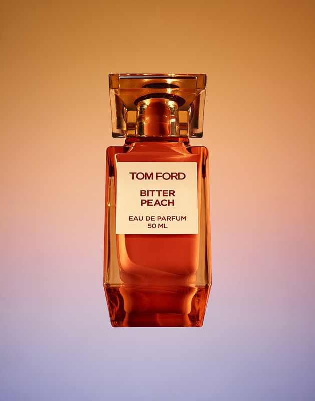 Tom Ford Bitter Peach perfume bottle. The transparent glass bottle has a amber liquid inside and a clear geometric stopper.